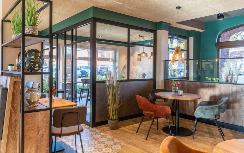 Modern gestalteter Café-Innenraum mit gemütlicher Möblierung, Glaswänden mit schwarzem Rahmen, runden Holztischen und gepolsterten Stühlen in Erdtönen; dekoriert mit Pflanzen, Vasen und stilvoller Hängelampe.