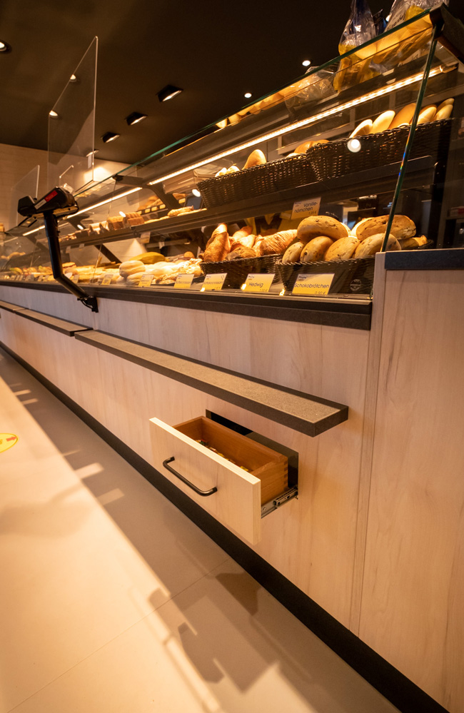 Das Bild zeigt eine Bäckereitheke mit diversen Backwaren in der Auslage und einer geöffneten Schublade im unteren Bereich.