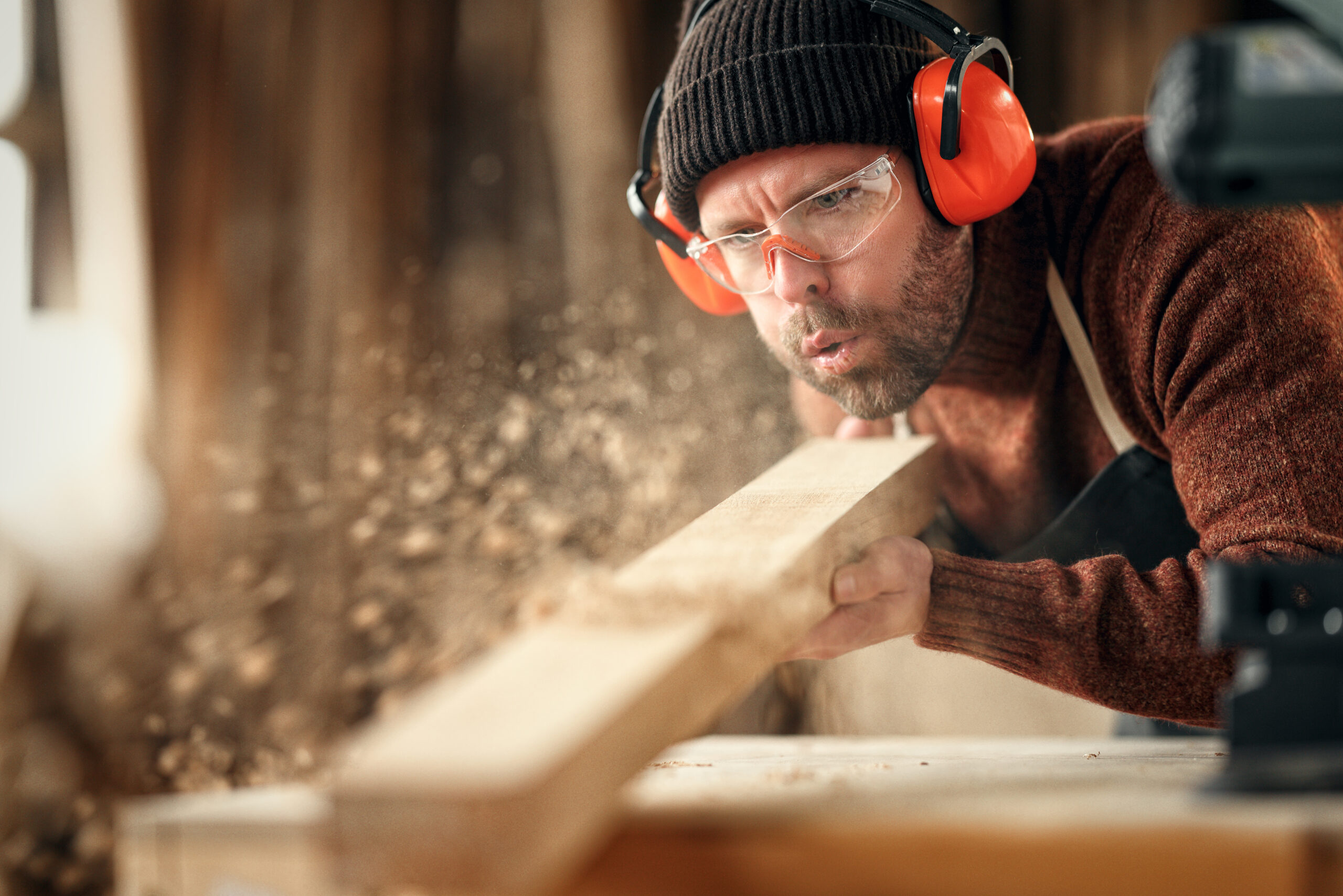 Handwerker bläst Holzstaub von einem frisch bearbeiteten Brett in einer Werkstatt.