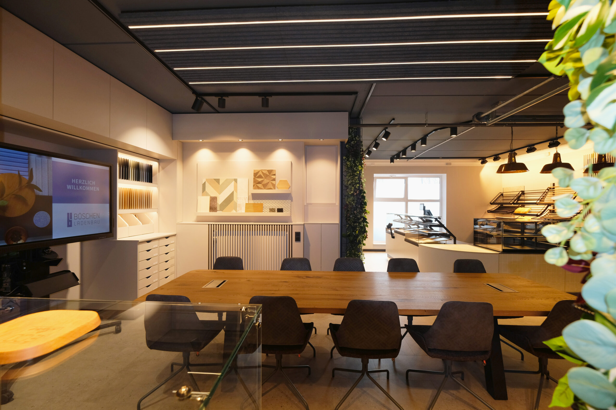 Moderner Showroom mit Besprechungstisch, Präsentationsbereich für Materialien, Monitor und angrenzendem Bäckereiverkaufsbereich.