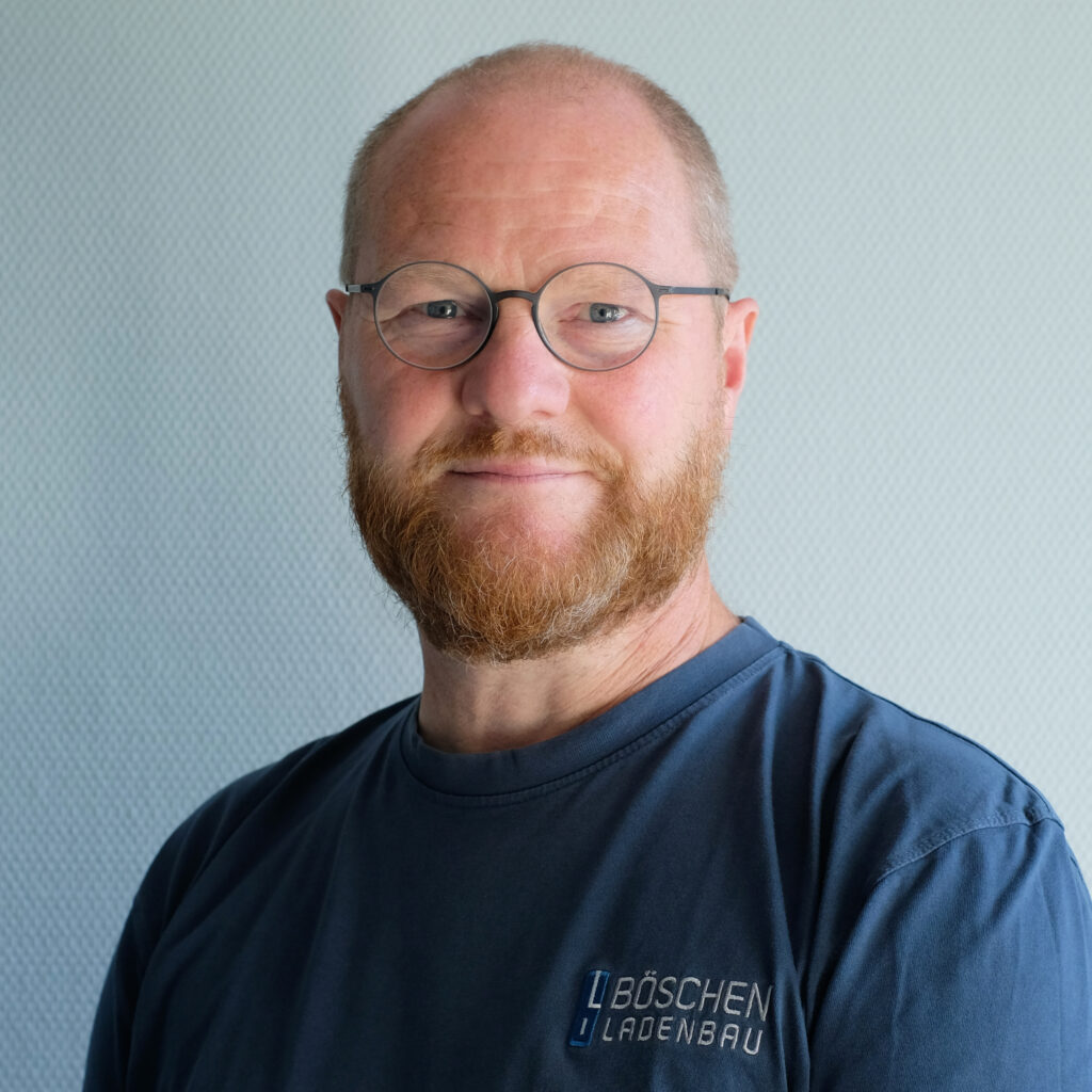 Porträt von Heino Ruschmeyer in dunkelblauem T-Shirt mit Bö­schen Ladenbau-Logo, vor hellem strukturiertem Hintergrund.