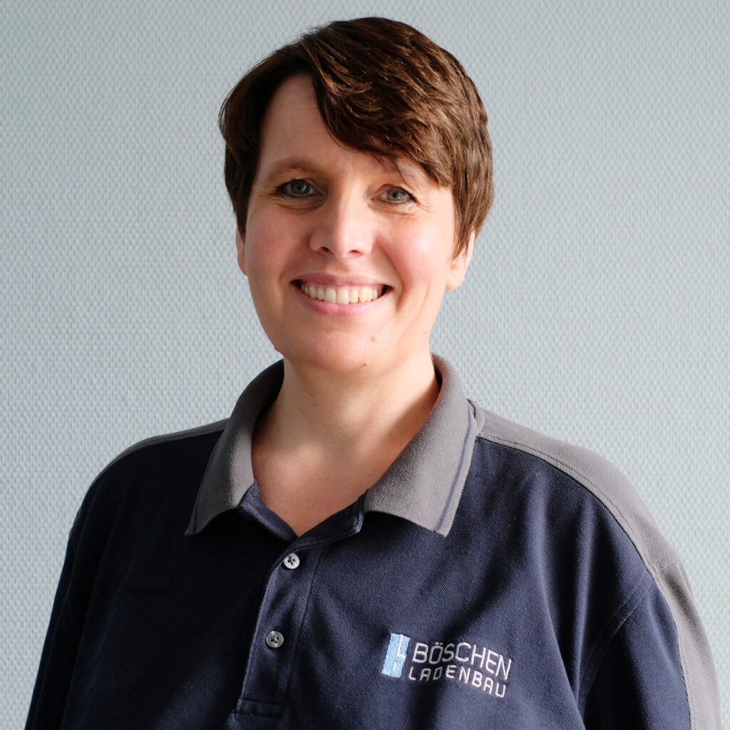 Porträt von Andrea Rasmussen in dunkelblauem T-Shirt mit Bö­schen Ladenbau-Logo, vor hellem strukturiertem Hintergrund.
