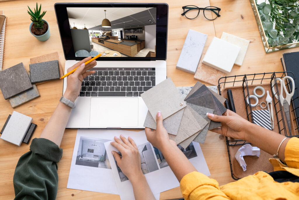 Zwei Mitarbeiter planen eine Innenraumgestaltung am Laptop mit 3D-Visualisierung eines Bäckereibereichs, Musterplatten und ausgedruckten Entwürfen auf dem Tisch.