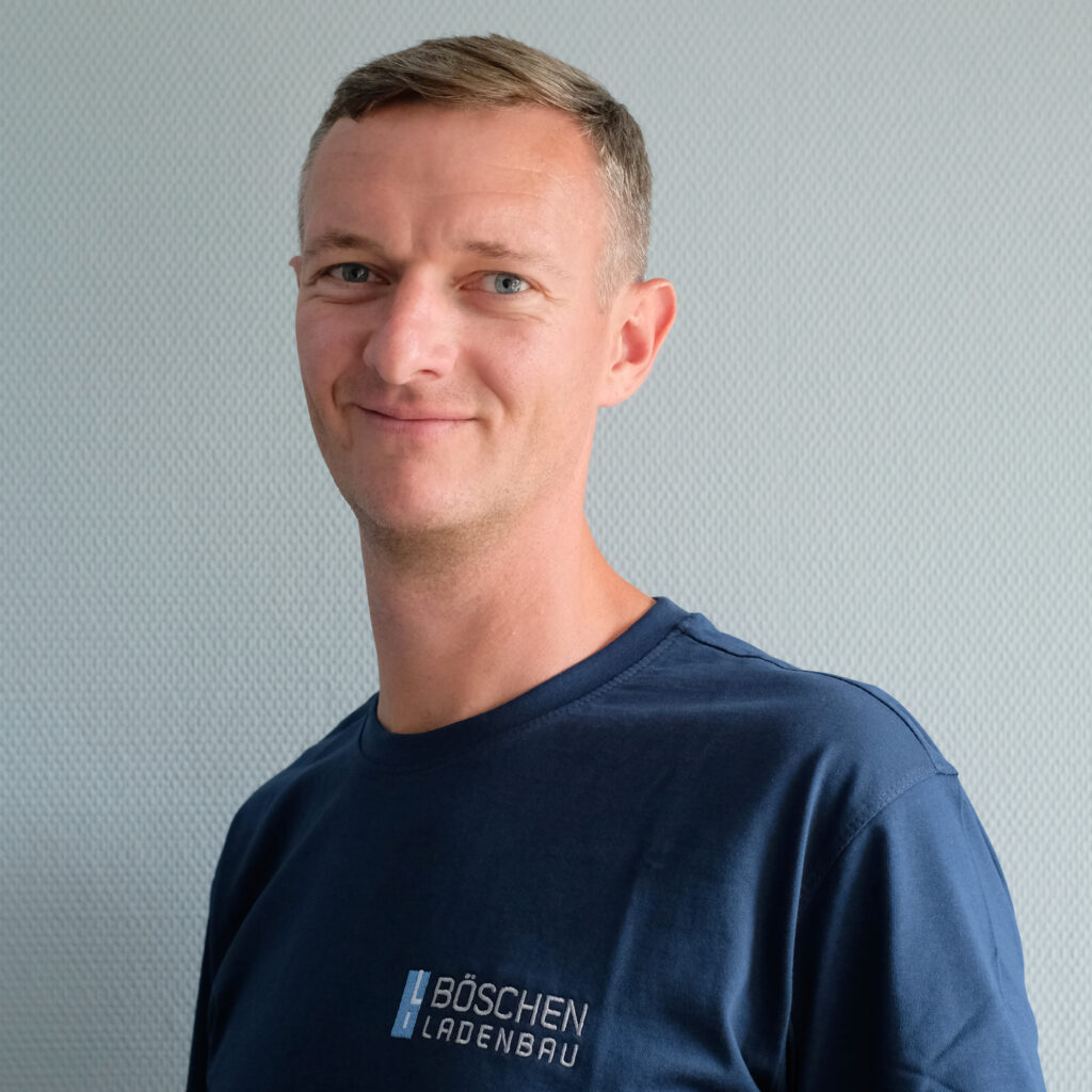 Porträt von Dima Kriger in dunkelblauem T-Shirt mit Bö­schen Ladenbau-Logo, vor hellem strukturiertem Hintergrund.