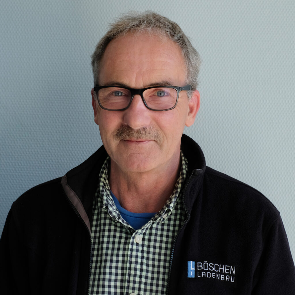 Porträt von Gisbert Greif in dunkelblauem T-Shirt mit Bö­schen Ladenbau-Logo, vor hellem strukturiertem Hintergrund.