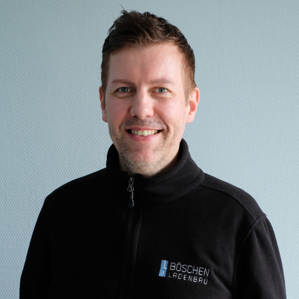 Porträt von Holger Tabeling in dunkelblauem T-Shirt mit Bö­schen Ladenbau-Logo, vor hellem strukturiertem Hintergrund.