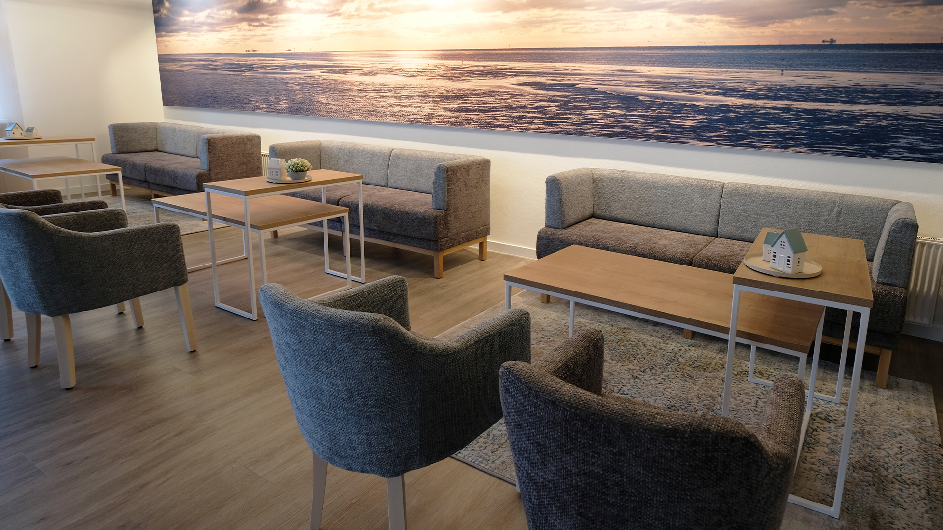 Sitzbereich eines Mitarbeiter-Pausenraums für eine Sparkasse in Cuxhaven mit gemütlichen grauen Sesseln und Sofas und kleinen Beistelltischen aus Holz und weißen Metall, welche individuell hingestellt werden können. Im Hintergrund ist ein riesiges Bild vom Sonnenaufgang über dem Meer zu sehen um die Verbindung zu Cuxhaven nochmal zu verdeutlichen.