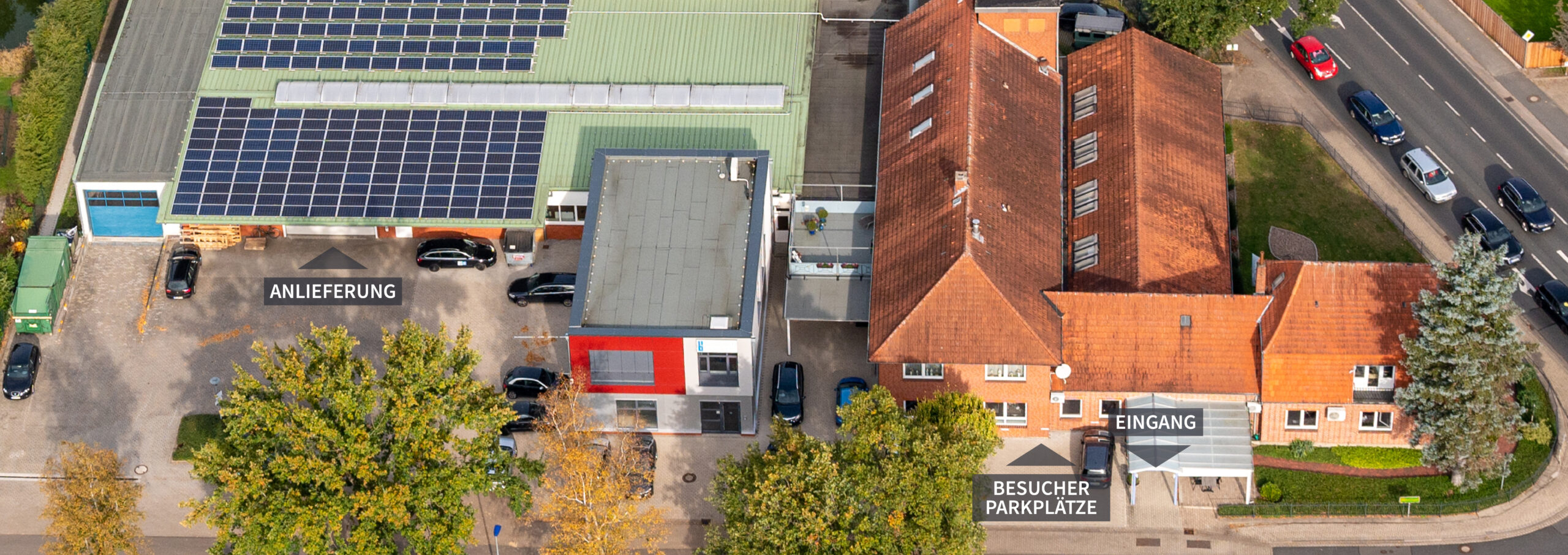 Luftaufnahme der Firma Böschen ladenbau GmbH in Ottersberg mit Produktionshalle, Solarzellen auf dem Dach, Bürogebäude und angrenzender Straße. Ebenso gibt es eine Beschriftung wo der Eingang, die Besucherparkplätze und die Anlieferung ist.