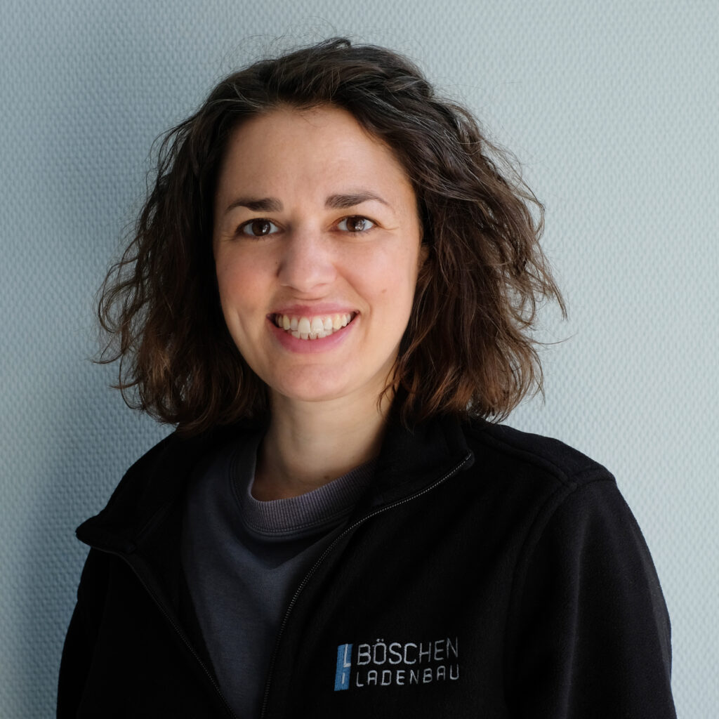 Porträt von Lisa Ehehalt in dunkelblauem T-Shirt mit Bö­schen Ladenbau-Logo, vor hellem strukturiertem Hintergrund.