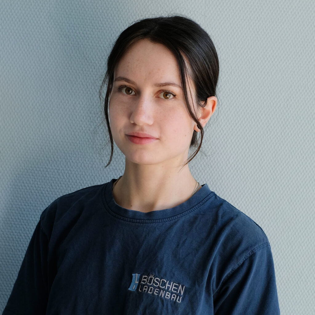 Porträt von Luci Schütt in dunkelblauem T-Shirt mit Bö­schen Ladenbau-Logo, vor hellem strukturiertem Hintergrund.