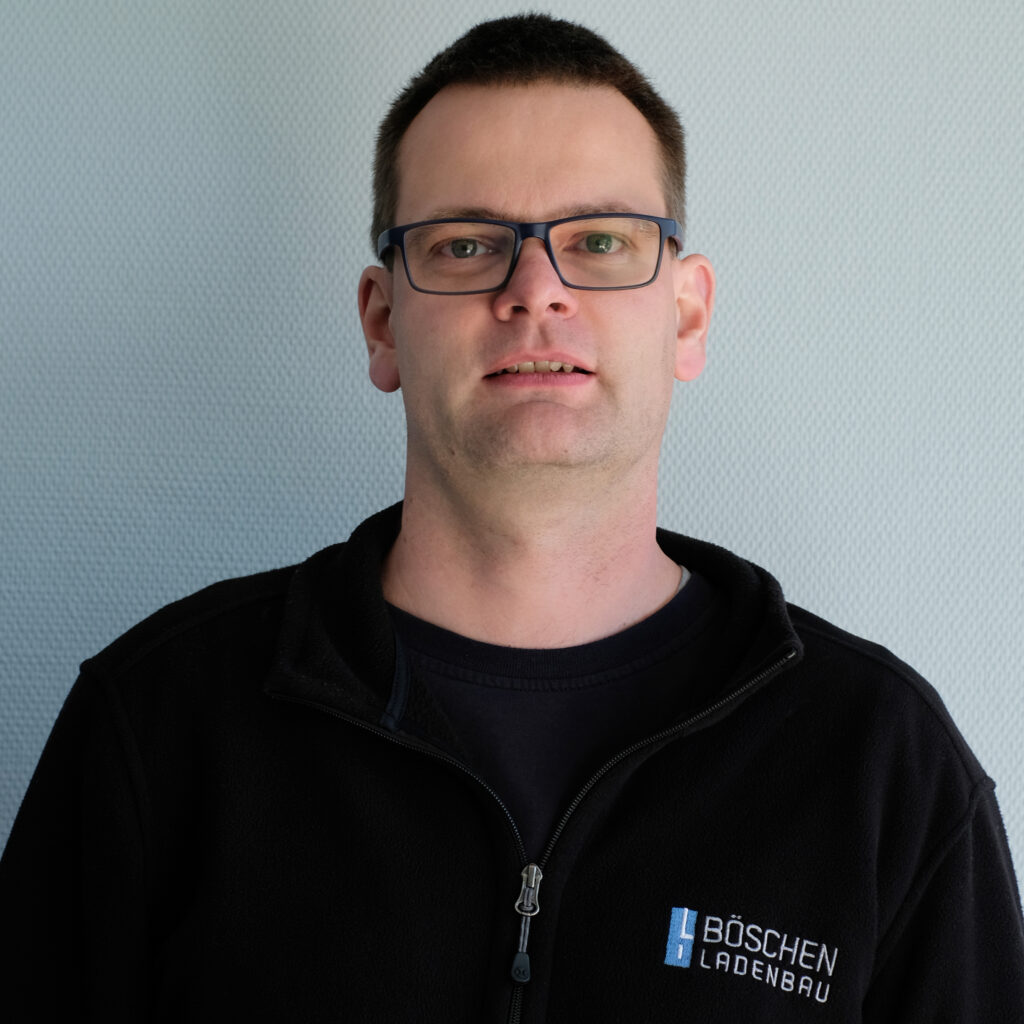 Porträt von Michael Hinkelmann in dunkelblauem T-Shirt mit Bö­schen Ladenbau-Logo, vor hellem strukturiertem Hintergrund.