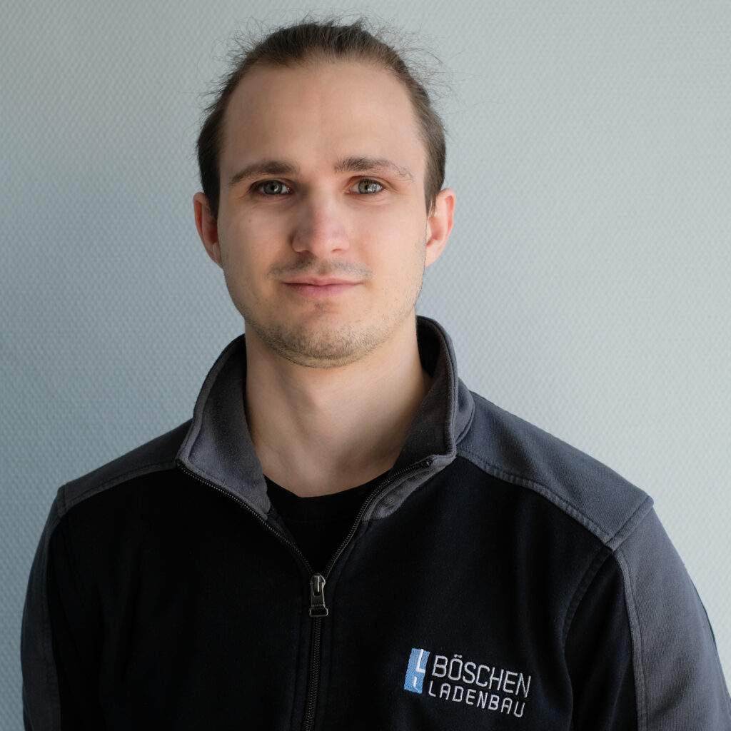 Porträt von Max-Luca Malende in dunkelblauem T-Shirt mit Bö­schen Ladenbau-Logo, vor hellem strukturiertem Hintergrund.