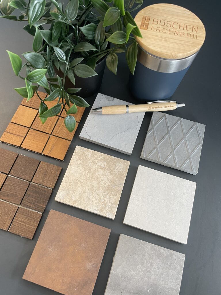 Materialcollage von verschiedenen Wandplatten und Hölzern mit einem Böschen Ladenbau Thermobecher und einem Kugelschreiber mit Logo sowie einer Kunstpflanze.