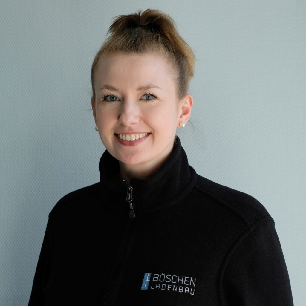 Porträt von Natalie Propst in dunkelblauem T-Shirt mit Bö­schen Ladenbau-Logo, vor hellem strukturiertem Hintergrund.