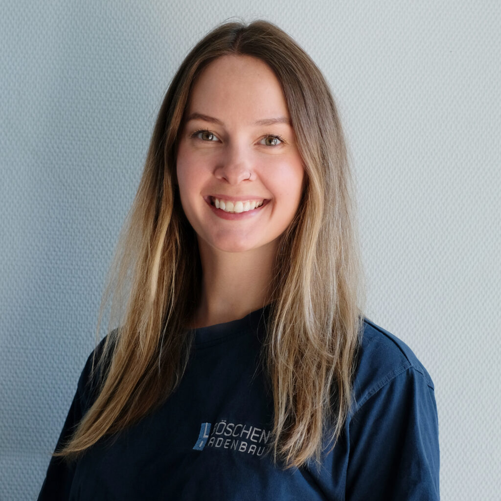 Porträt von Lia Fastenau in dunkelblauem T-Shirt mit Bö­schen Ladenbau-Logo, vor hellem strukturiertem Hintergrund.