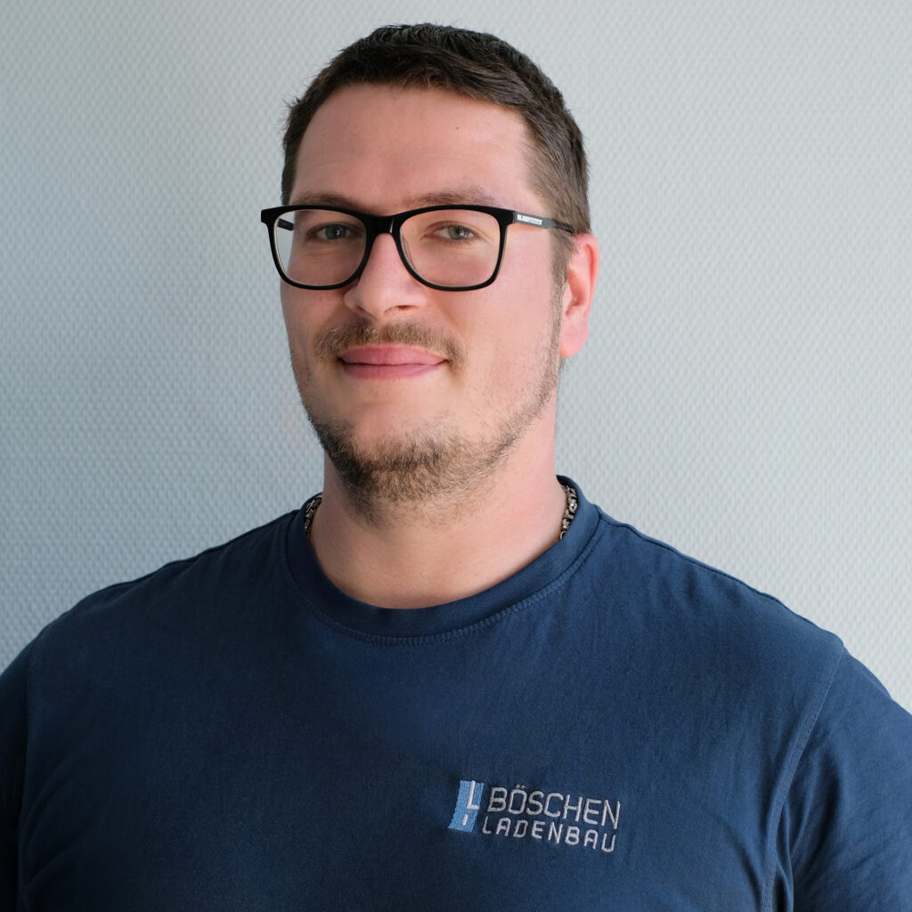 Porträt von Sascha Riedel in dunkelblauem T-Shirt mit Bö­schen Ladenbau-Logo, vor hellem strukturiertem Hintergrund.