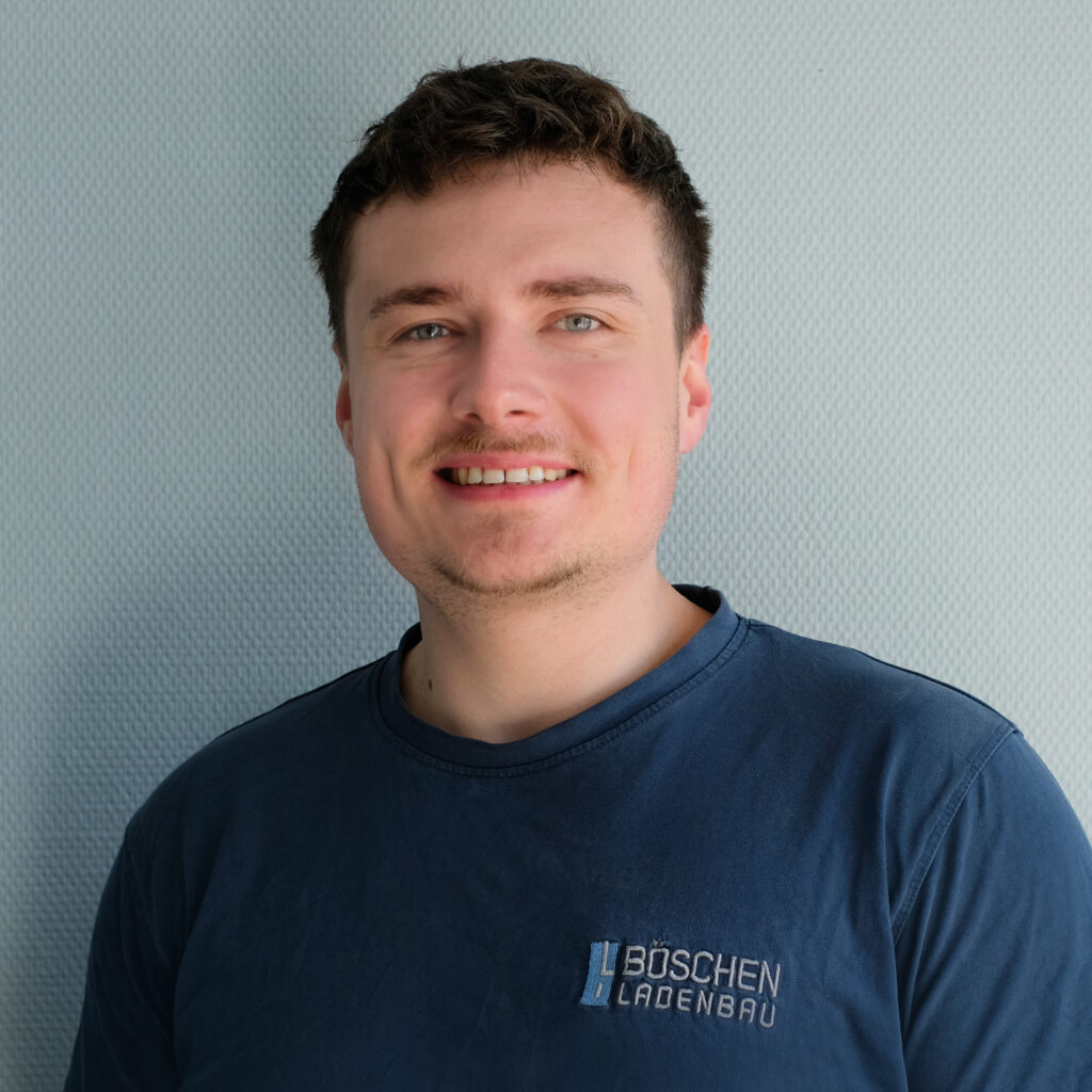 Porträt von Sean Wundes in dunkelblauem T-Shirt mit Bö­schen Ladenbau-Logo, vor hellem strukturiertem Hintergrund.