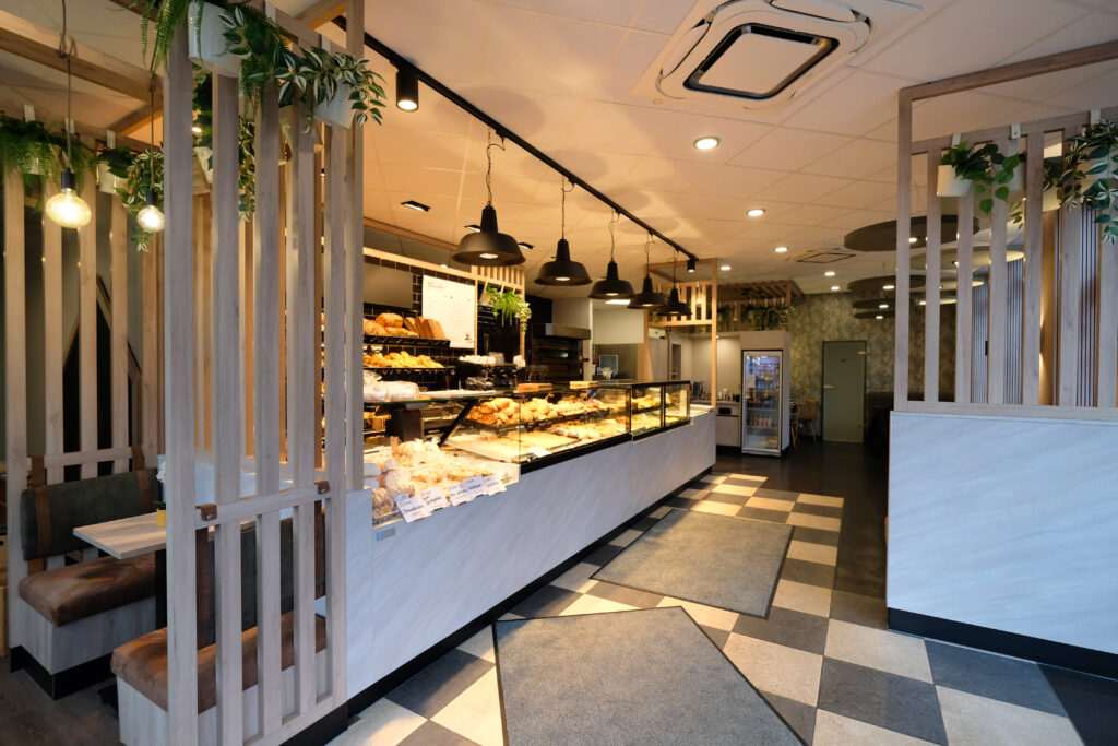 Ansicht weißer heller schöner Bäckereitresen mit schwarzen Hängeleuchten und Strahlern versehen. Volles übersichtliches Brotregal mit einem weiß-schwarzem Kachelboden und Lamellen-Trennelementen für den Sitzbereich.