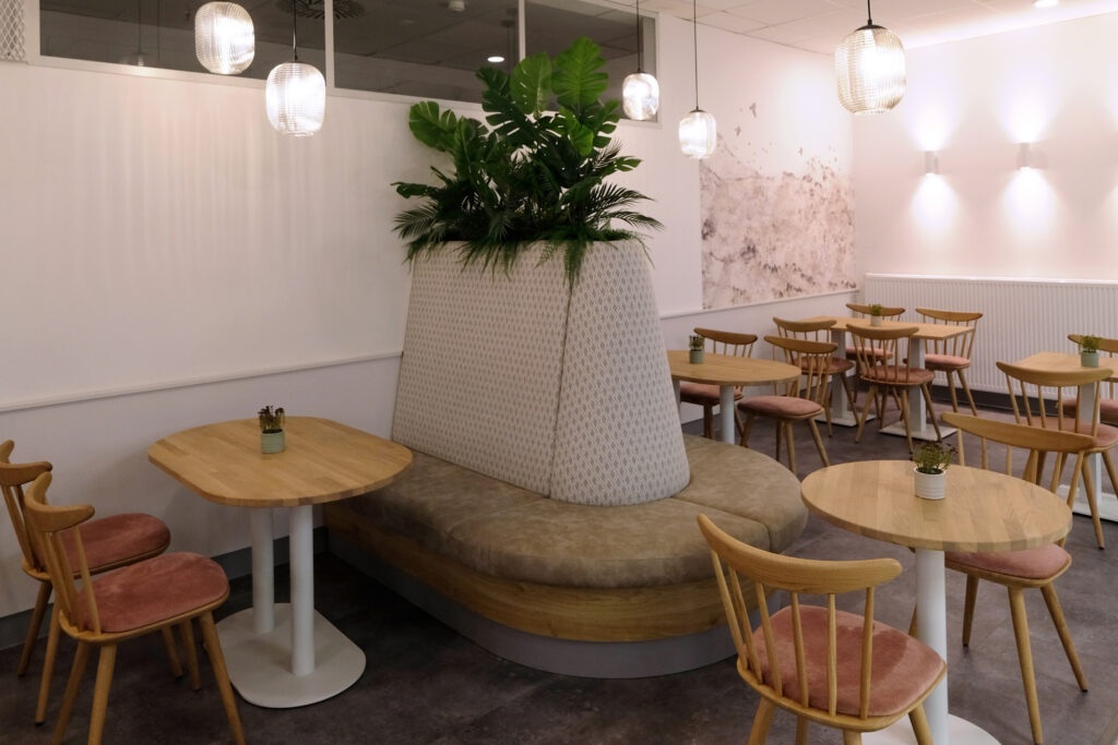 Moderner heller Sitzbereich in hellen weiß, beige, braun und Rosetönen mit einer tollen Wandtapete und Hängeleuchten aus Glas. Die Tischgestelle bestehen aus einer Holzplatte und einem weißen Fußgestell aus Metall. In der Mitte ist eine maßgefertigte Sitzbank zu sehen in der im oberen bereich Pflanzen eingearbeitet wurden, welche direkt als Sichtschutz dienen.
