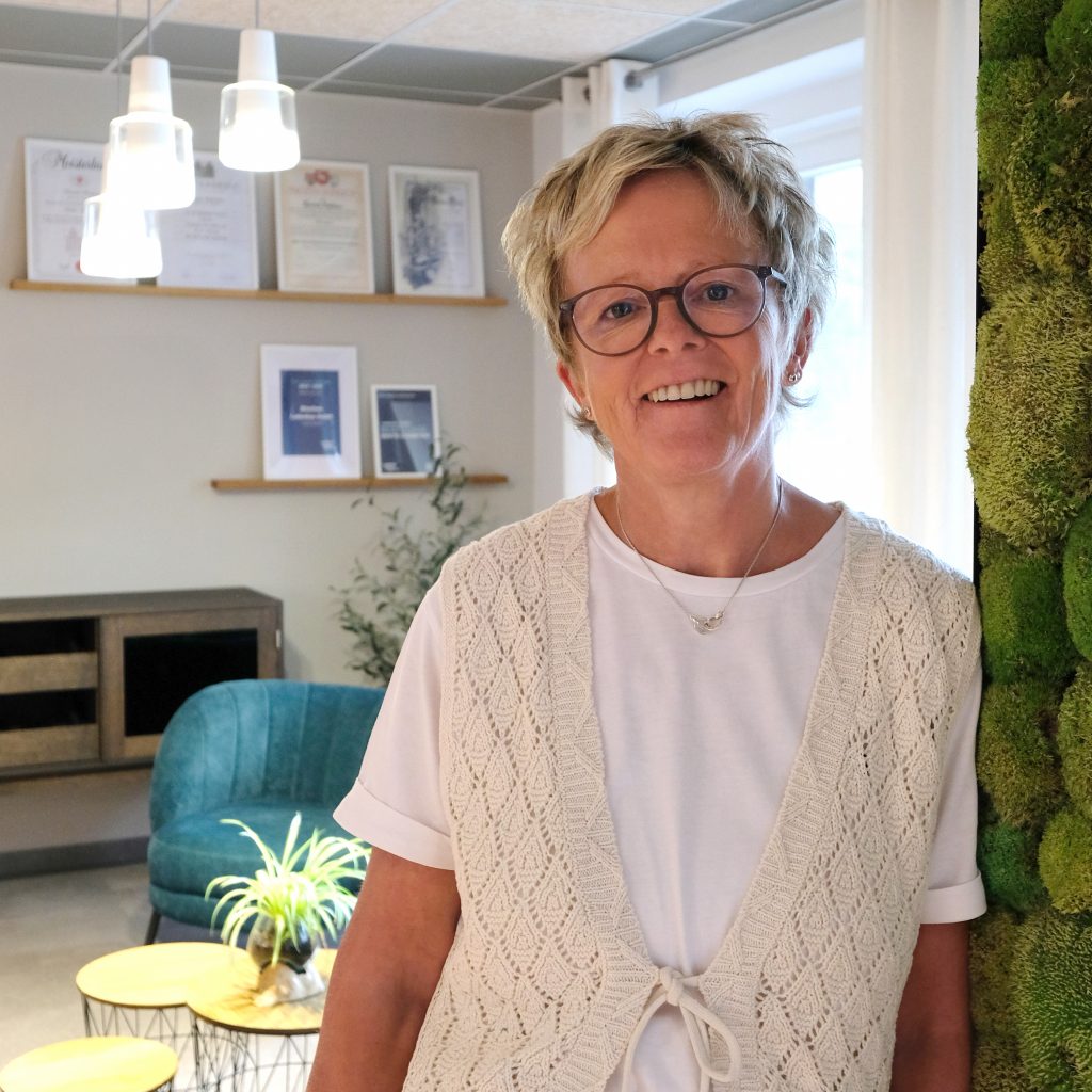 Porträt von Elke Böschen im weißen T-Shirt mit weißer Strickweste darüber steht in dem modern gestalteten Empfangsbereich von Böschen Ladenbau.